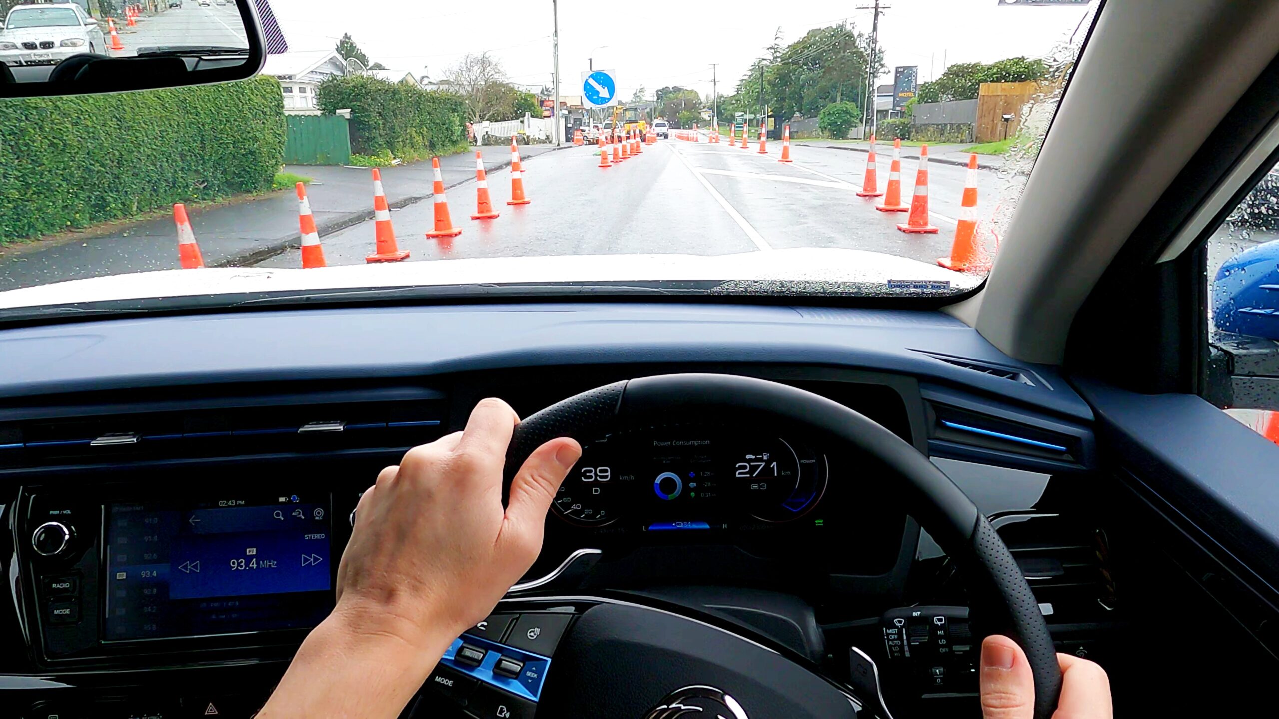 A view of the steering wheel and dashboard taken while driving on a suburban street, showing my hands on the wheel and the road ahead